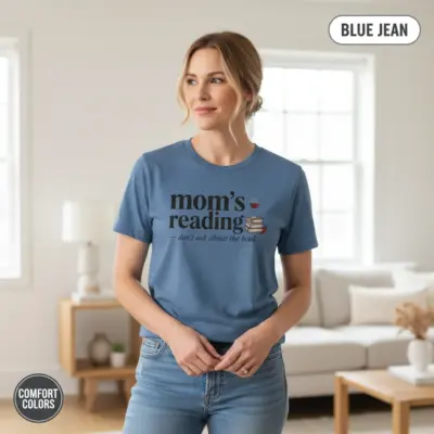 Blue Jean Comfort Colors garment-dyed tee printed with "Mom's Reading Don't Ask About the Book" funny bookish typography, worn by a woman indoors in casual setting